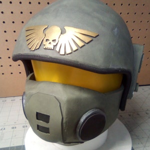 May include: A green and gold helmet with a skull and wings design. The helmet has a black visor and a black face mask with a circular opening.