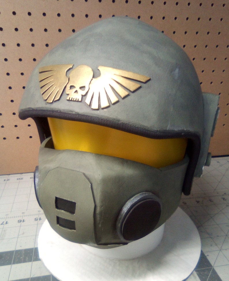 May include: A green and gold helmet with a skull and wings design. The helmet has a black visor and a black face mask with a circular opening.