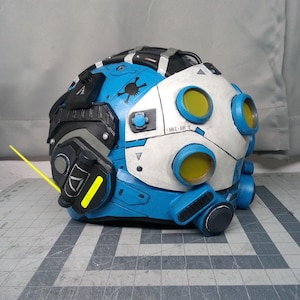 Op de afbeelding: Een blauwe, witte en zwarte helm met een ontwerp met meerdere lenzen en gele ronde lenzen. De helm bevat een zwart schedelpictogram, een dunne gele antenne en diverse mechanische details. Het witte voorpaneel toont de tekst 68Z-E21.