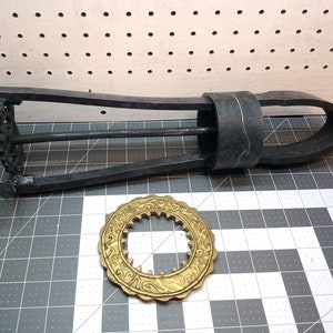 May include: A black metal contraption with a gear mechanism and a black rubber band. A gold metal decorative piece with a circular design is also pictured.