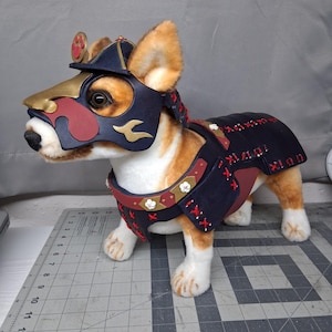 May include: A plush corgi dog toy wearing a dark blue and red samurai-style armor set. The armor includes a headpiece with gold accents and a body harness featuring red lacing, small white flower details, and gold trim over the back and chest.