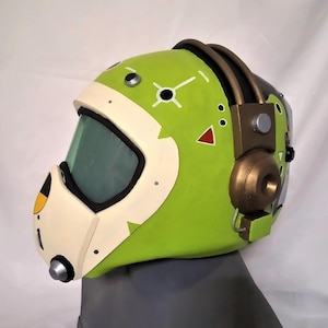 Könnte beinhalten: Ein grün-weißer Helm mit goldenem Headset und klarem Visier. Der Helm hat verschiedene schwarze und rote Markierungen.