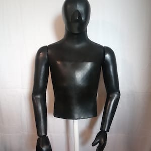 Puede incluir: Un torso de maniquí negro con brazos y manos articulados. El maniquí tiene un acabado liso y mate y está montado sobre un poste blanco. La cabeza no tiene rasgos, con una nariz y una zona de ojos definidas.