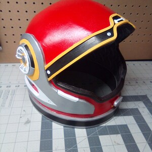 May include: A red, black, and yellow helmet with a silver visor. The helmet has a circular design on the side and a black band around the top.