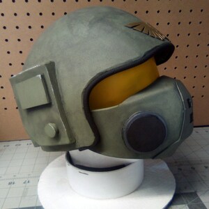 May include: A green and brown helmet with a yellow visor and a gold emblem. The helmet has a black circular detail on the side.