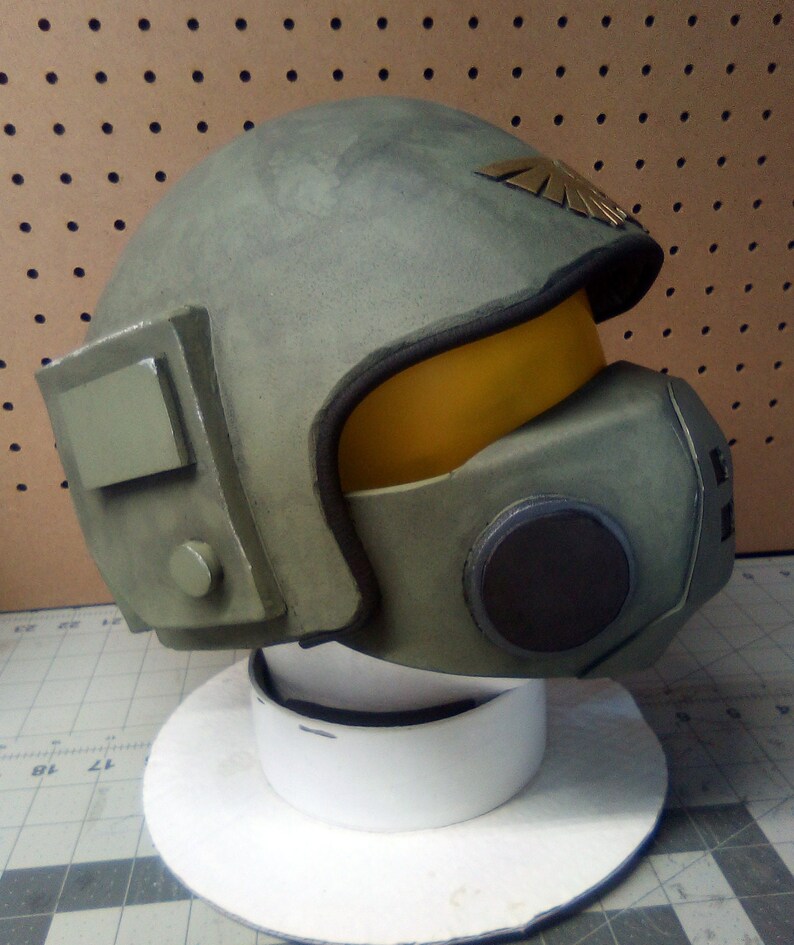 May include: A green and brown helmet with a yellow visor and a gold emblem. The helmet has a black circular detail on the side.