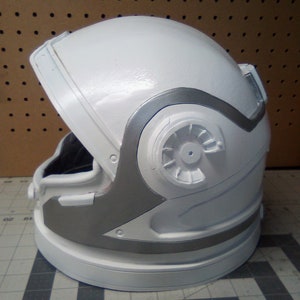 May include: A white astronaut helmet with a silver trim around the visor and a silver detail on the side. The helmet is made of a hard plastic material.