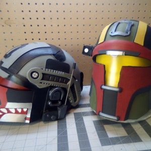 Könnte beinhalten: Zwei Helme, einer grau und schwarz mit einem roten und weißen Streifen, der andere grün, gelb und rot mit einem schwarzen Streifen. Beide Helme haben ein Visier und eine kleine Antenne.