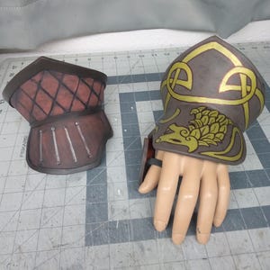 Könnte beinhalten: Zwei mittelalterliche Panzerhandschuhe. Einer ist braun mit einem roten Rautenmuster, der andere ist grau mit goldenen keltischen Knotenmustern und einem Drachen. Der Panzerhandschuh befindet sich auf einer Schaufensterpuppenhand.