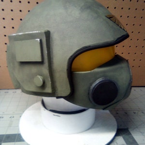 May include: A green and black helmet with a yellow visor and a black circular detail on the side. The helmet is on a white display stand.