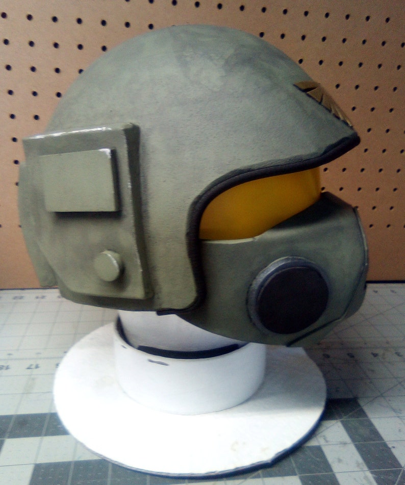 May include: A green and black helmet with a yellow visor and a black circular detail on the side. The helmet is on a white display stand.