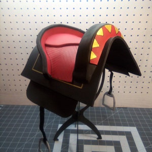 May include: A black leather saddle with a red padded seat and gold accents. The saddle is mounted on a black metal stand.