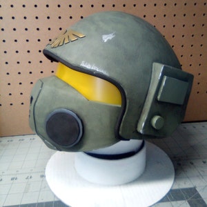 May include: A green and yellow helmet with a black visor and a gold eagle emblem. The helmet is made of a hard plastic material and has a textured surface.