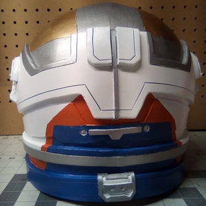 May include: A white, silver, and gold helmet with orange and blue accents. The helmet has a silver band around the bottom and a silver buckle on the front.