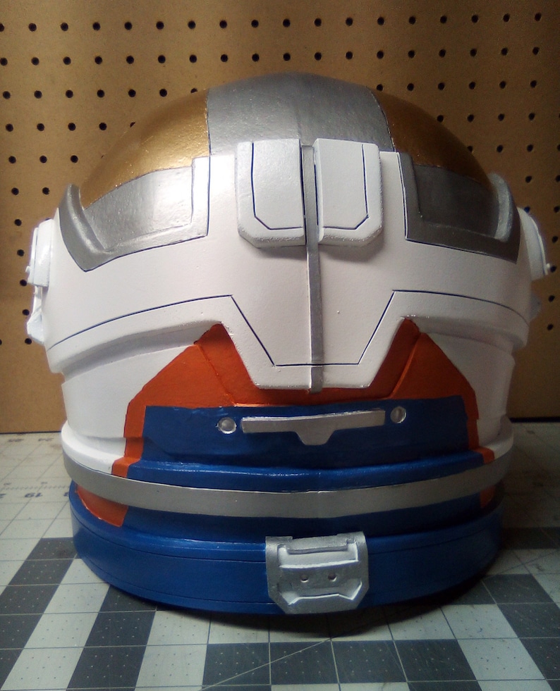 May include: A white, silver, and gold helmet with orange and blue accents. The helmet has a silver band around the bottom and a silver buckle on the front.