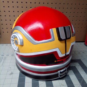May include: A red, yellow, and silver helmet with a black visor. The helmet has a circular design on the side and a black band around the bottom.