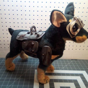 Könnte beinhalten: Ein Plüschhundespielzeug mit schwarzem und hellbraunem Fell, das eine Metallbrille und einen Sattel trägt. Die Ohren des Hundes sind hellbraun, und seine Pfoten sind hellbraun. Die Metallaccessoires haben ein gealtertes Aussehen.