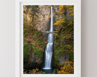 May include: A framed photograph of a waterfall cascading down a rocky cliff. A small bridge spans the water. The surrounding foliage displays autumn colors of yellow, orange, and green.