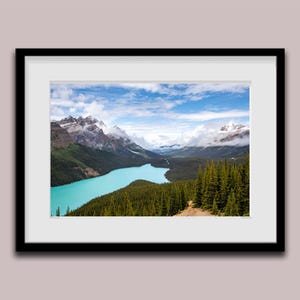 Peut inclure: Tirage encadré d'un paysage avec un lac turquoise, des forêts vertes et des montagnes enneigées sous un ciel bleu nuageux. L'impression est dans un cadre noir avec un passe-partout blanc.