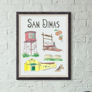 May include: A watercolor illustration of San Dimas, California, featuring a water tower, a train, a building, and a horse. The illustration is in a brown frame and is hanging on a white brick wall.