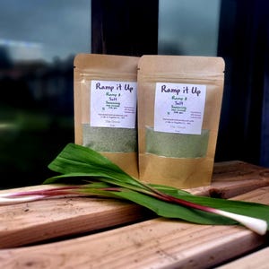 May include: Two brown paper bags with white labels that say "Ramp it Up Ramp & Salt Seasoning Fine Ground" and "Ramp it Up Ramp & Salt Seasoning Fine Ground". The bags are sitting on a wooden surface with a green plant in front of them.
