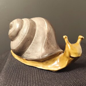 May include: A ceramic snail figurine featuring a glossy, mustard-yellow body and a textured, matte-finish shell in shades of dark gray and brown. The shell displays a spiral pattern with fine, etched lines, while the head has two distinct eye stalks.