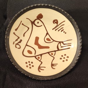 May include: A round ceramic plate with a cream-colored center and a dark brown rim. The plate features a brown abstract design of a creature with stylized features. The rim has a textured, ridged pattern.