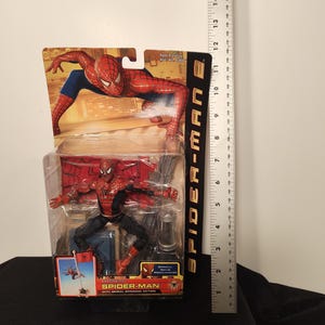May include: Spider-Man 2 action figure in packaging. The figure is red and black, with blue accents. The packaging features Spider-Man in action poses. The figure is approximately 6 inches tall.