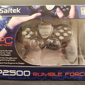 May include: A Saitek P2500 Rumble Force PC gamepad in its original retail packaging. The black controller features silver buttons and a central silver panel. The box displays the product name, features, and a graphic of the device in use.