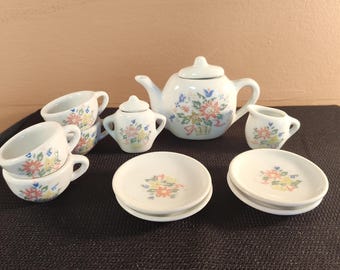 May include: A miniature white porcelain tea set with floral designs. The set includes a teapot, sugar bowl, creamer, cups, and saucers. The floral pattern features pink, yellow, and blue flowers.