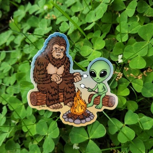 May include: A sticker featuring a cartoon Bigfoot and an alien roasting marshmallows over a campfire. The Bigfoot is brown and furry, while the alien is green with large eyes. The campfire is orange and yellow.