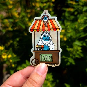 May include: A whimsical sticker featuring a cartoon yeti selling ice cream from a brown cart. The cart has a striped orange and yellow awning and the text "I SCREAM". The yeti has blue fur and is holding three ice cream cones.