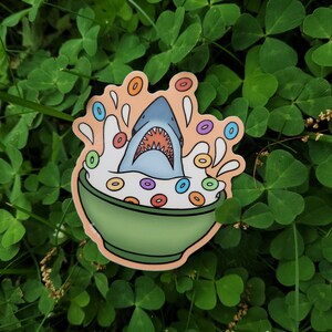 May include: A sticker of a blue shark swimming in a bowl of milk with colorful cereal rings. The shark has an open mouth with sharp teeth. The bowl is green and has a white rim.