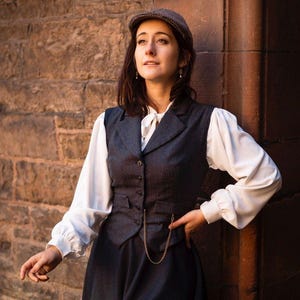 Könnte beinhalten: Ein Outfit im Vintage-Stil mit einer grauen Tweedweste mit goldener Kette, einer weißen Bluse und einem dunkelblauen Rock. Abgerundet wird das Ensemble durch eine passende Tweedkappe. Das Outfit erinnert an eine klassische, historische Ästhetik.