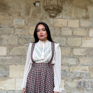 Op de afbeelding: Een vrouw in een vintage outfit. De outfit bestaat uit een witte blouse met polkadots en een hoog getailleerde geruite rok in rood, zwart en wit, met bretels. De achtergrond is een stenen muur.
