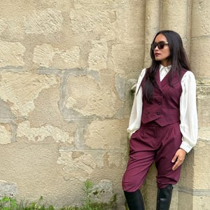 Può includere: Un gilet bordeaux e pantaloni abbinati al ginocchio sono abbinati a una camicetta bianca e stivali neri. L'outfit è indossato con occhiali da sole. L'ensemble è ambientato contro un muro di pietra strutturato e fogliame verde.