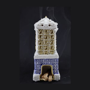 May include: White ceramic incense burner shaped like a fireplace with gold and blue details. The burner has a small opening at the bottom where incense sticks can be placed. Smoke is rising from the top of the burner.