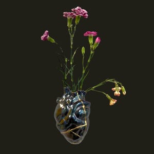 May include: A unique, heart-shaped vase with a dark blue and gold finish, holding pink carnations. The vase has visible gold accents, and the flowers add a pop of color against the dark background. A decorative floral arrangement.