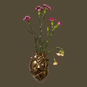 May include: A bronze-colored heart-shaped vase with gold accents, holding several stems of pink and purple carnations. The vase has a unique, textured surface and a kintsugi-inspired design.