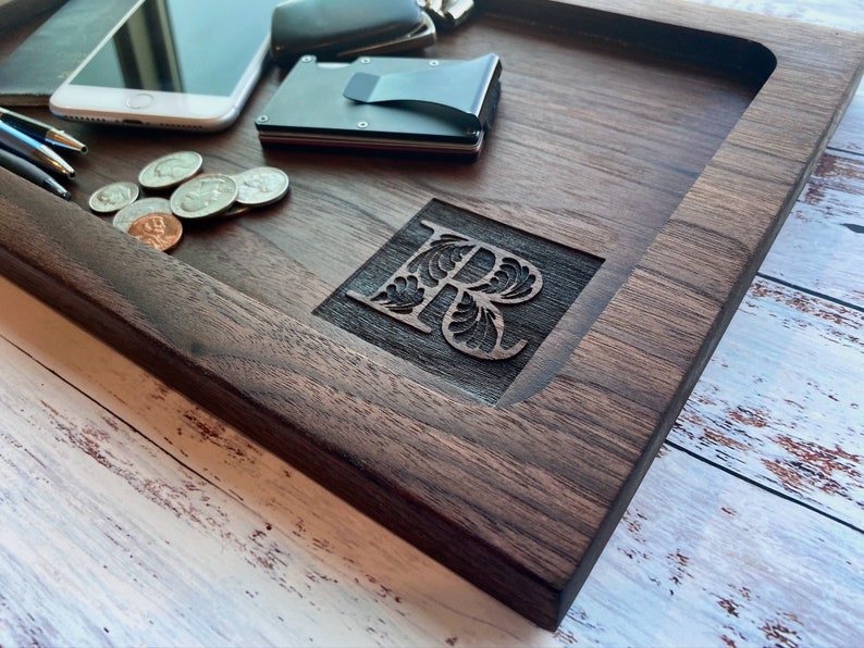 Pu&ograve; includere: Un vassoio in legno di noce scuro con una lettera "R" intagliata. Il vassoio rettangolare ha bordi rialzati. Monete, penne, un telefono e un portafoglio sono visibili sul vassoio. Il vassoio misura circa 30 cm di lunghezza.