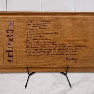 May include: A wooden cutting board with a recipe for Aunt B's Mac & Cheese. The recipe is handwritten in black ink and includes ingredients like elbow macaroni, shredded cheese, and butter. The board is displayed on a black metal stand.