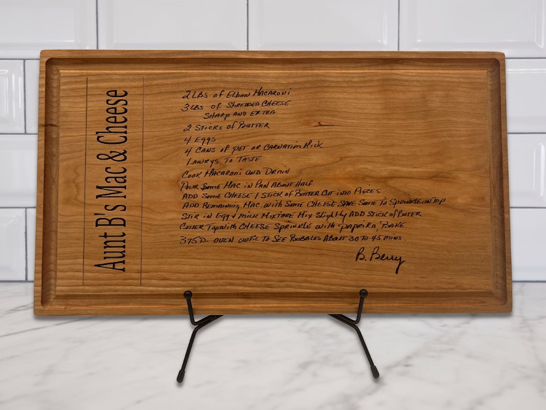 May include: A wooden cutting board with a recipe for Aunt B's Mac & Cheese. The recipe is handwritten in black ink and includes ingredients like elbow macaroni, shredded cheese, and butter. The board is displayed on a black metal stand.