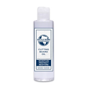 Pode incluir: Uma garrafa transparente de &oacute;leo para t&aacute;buas de corte da Battle Born Wood Shop. O r&oacute;tulo apresenta um design azul e branco com as palavras "Cutting Board Oil", "Maintain, Protect, Restore" e "Food Safe". A garrafa tem cerca de 13 cm de altura.