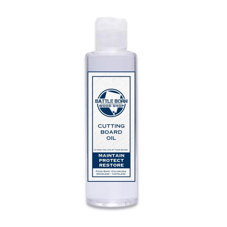 Pode incluir: Uma garrafa transparente de &oacute;leo para t&aacute;buas de corte da Battle Born Wood Shop. O r&oacute;tulo apresenta um design azul e branco com as palavras "Cutting Board Oil", "Maintain, Protect, Restore" e "Food Safe". A garrafa tem cerca de 13 cm de altura.