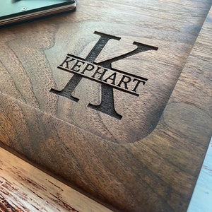 Pode incluir: Uma t&aacute;bua de corte de madeira castanha escura com o nome "KEPHART" gravado no centro. A letra "K" &eacute; exibida de forma proeminente acima do nome. Uma pequena carteira e moedas s&atilde;o vis&iacute;veis no canto superior esquerdo.