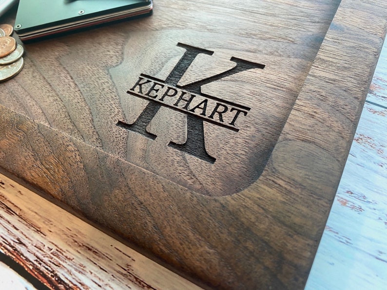 Pode incluir: Uma t&aacute;bua de corte de madeira castanha escura com o nome "KEPHART" gravado no centro. A letra "K" &eacute; exibida de forma proeminente acima do nome. Uma pequena carteira e moedas s&atilde;o vis&iacute;veis no canto superior esquerdo.
