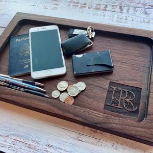 Pu&ograve; includere: Un vassoio in legno scuro con interno incassato contiene un passaporto, uno smartphone, penne, monete, un portafoglio e chiavi. Il vassoio presenta un'iniziale scolpita e un design rettangolare liscio.