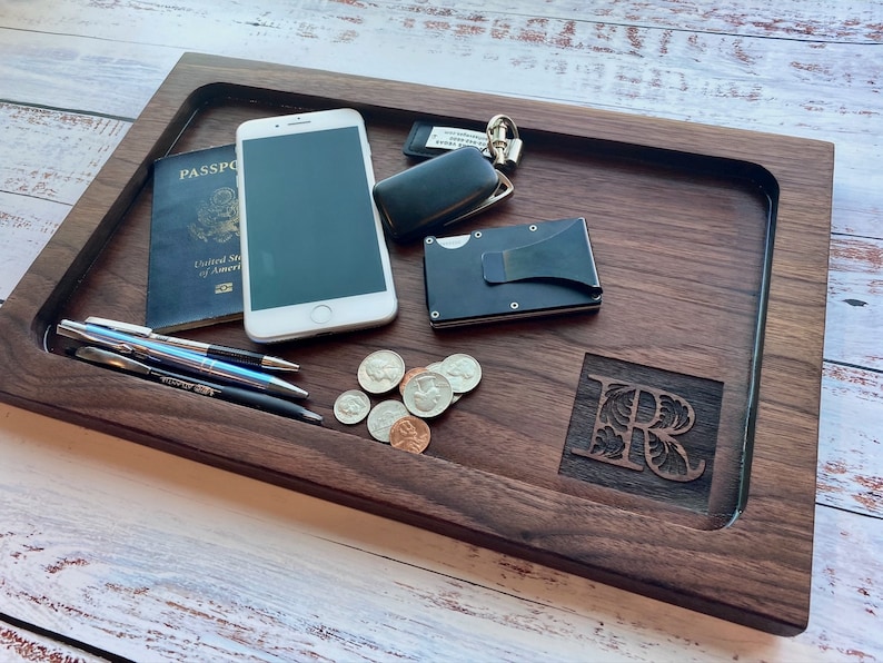 Pu&ograve; includere: Un vassoio in legno scuro con interno incassato contiene un passaporto, uno smartphone, penne, monete, un portafoglio e chiavi. Il vassoio presenta un'iniziale scolpita e un design rettangolare liscio.