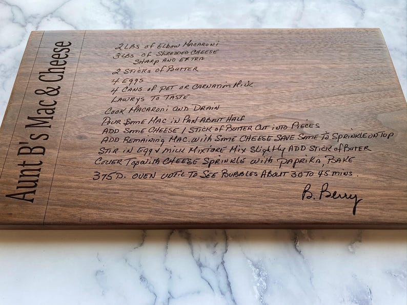 May include: A rectangular wooden cutting board with the text "Aunt B's Mac & Cheese" engraved on the side. The board features a handwritten recipe for mac and cheese, including ingredients and instructions.