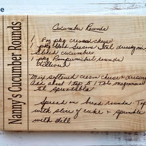 May include: A rectangular maple cutting board with a handwritten recipe for "Nanny's Cucumber Rounds" engraved on it. The recipe includes ingredients like cream cheese, Italian dressing, and dill. The text "Maple" is in the upper left corner.